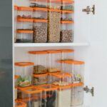Neatly organized pantry shelves featuring diverse grains and seeds in transparent containers with orange lids.